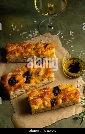 Freshly baked homemade focaccia topped with garlic and olives served on parchment paper. Accompanied by olive oil dip and a glass of white wine on a t Stock Photo