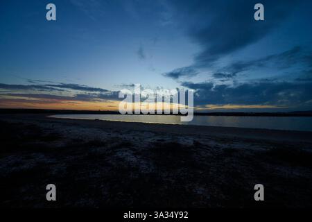 A peaceful lake view at dusk with vibrant skies in lonely field. The calm water reflects the blue and orange hues from the sky. Silhouettes of distant Stock Photo