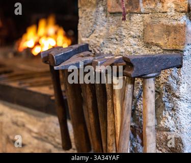 A row of well-worn blacksmith hammers with wooden handles rests against a rustic brick wall, illuminated by the warm glow of a forge fire in the backg Stock Photo