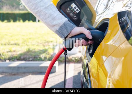 Close-up of a hand plugging a red cable into an electric car at a charging station, highlighting the eco-friendly transportation concept on a sunny da Stock Photo