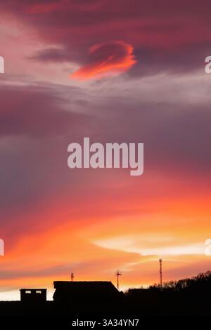 A vibrant sunset with dramatic hues of orange, pink, and purple fills the evening sky. Silhouettes of rooftops and antennas add contrast to the colorf Stock Photo
