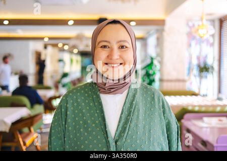Asian woman wearing a green hijab smiling warmly in a cozy cafe. Her serene expression reflects cultural beauty and traditional attire, emphasizing et Stock Photo
