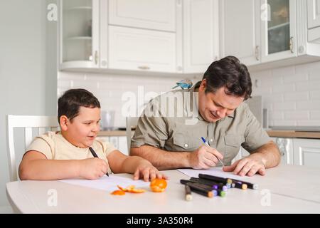 A father and son enjoy drawing together at the kitchen table, surrounded by art supplies and fresh fruit, while a small pet bird perches on the father Stock Photo