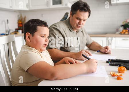 A father and son sit together at a kitchen table drawing, while a bird perches nearby. The atmosphere is warm and cozy, highlighting family bonding an Stock Photo