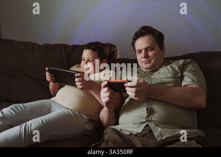 A father and son sit together on a cozy sofa, deeply engaged in playing video games. The image captures the essence of family bonding and shared leisu Stock Photo