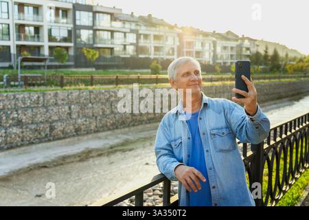 A senior man in casual clothing stands by a railing, taking a selfie with his smartphone. The background shows modern apartments and a sunny sky, capt Stock Photo