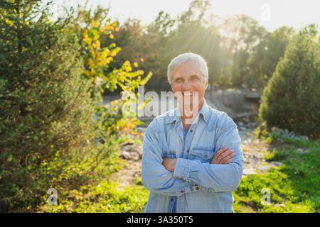 A cheerful senior man stands outdoors, arms crossed, in a sunlit park. He wears a denim shirt and smiles warmly, surrounded by greenery and natural li Stock Photo