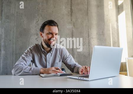 A professional man sits smiling at a desk, engaging with his laptop while taking notes. Natural light filters into the office, creating a productive a Stock Photo