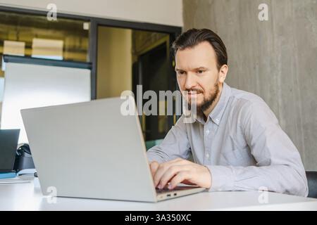 A man works on a laptop at his office desk, wearing a light shirt. The modern office includes a large window and a flip chart, suggesting a profession Stock Photo