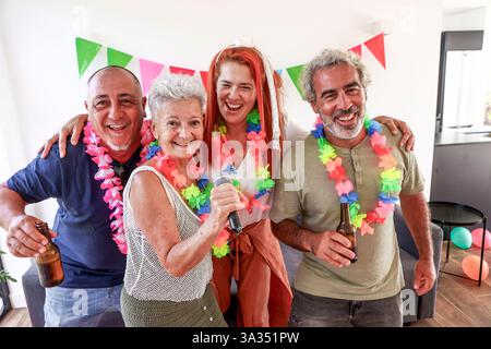 Elderly group of friends, dressed in colorful leis, celebrate at a vibrant over-50’s party, singing and having fun with drinks. Stock Photo