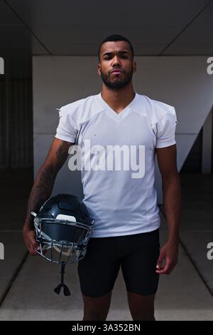 A focused American football player stands outdoors holding his helmet, wearing a white jersey. His determined expression embodies the spirit of the sp Stock Photo