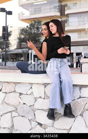 Young girlfriends sit on a stone wall, back to back, using smartphones. They wear casual outfits, enjoying a sunny day together in front of modern bui Stock Photo