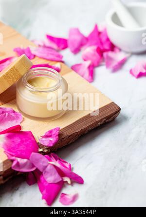 A glass jar of rose face cream with a wooden lid sits on a wooden board, surrounded by vibrant pink rose petals. A white mortar is in the background, Stock Photo