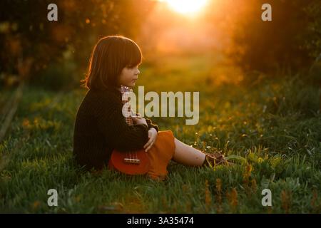 A child sits in a grassy field, holding a ukulele, with the warm glow of a sunset in the background. The scene captures a peaceful, reflective moment. Stock Photo