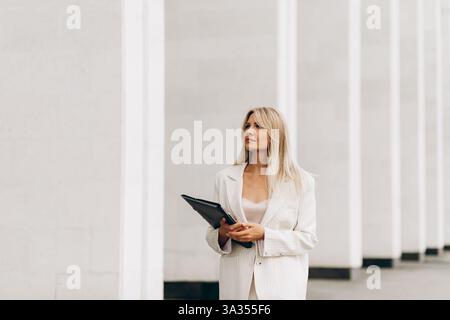 A professional woman holding a folder walks confidently beside a modern architectural structure. She is dressed in a stylish suit, exuding confidence Stock Photo