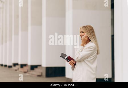 A businesswoman in a white suit stands outside a modern building, engaged in a phone conversation. She holds a book, exuding confidence and profession Stock Photo