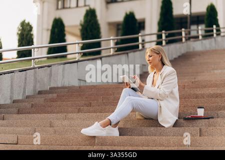 A businesswoman in casual attire sits on outdoor steps with a digital tablet, concentrating on her work. A takeaway coffee cup and papers lie beside h Stock Photo