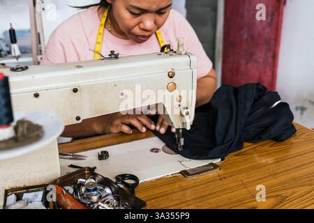 A asian seamstress works attentively with a vintage sewing machine, sewing black fabric in a creative workspace. Tools like spools and bobbins accompa Stock Photo