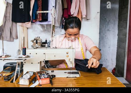 A asian seamstress diligently works with a sewing machine, surrounded by tools and fabrics in a cozy tailor shop. Her focus and precision highlight he Stock Photo