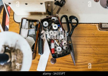 A collection of sewing tools and supplies, including scissors, spools, and a screwdriver, laid out on a wooden work surface, highlighting a well used Stock Photo