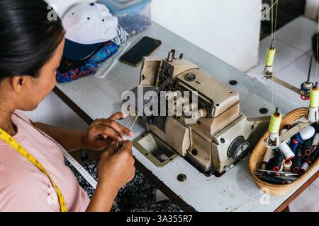 A asian seamstress operates a sewing machine in a textile workshop, focusing on detail. Nearby are spools of thread in a basket. The setting is an art Stock Photo
