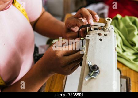 Close up of hands asian seamstress adjusting an industrial sewing machine in a workshop. A measuring tape is visible around the neck, and fabric lies Stock Photo