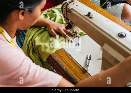 A asian seamstress skillfully sewing green fabric using a sewing machine. The workspace is equipped with essential tools like scissors and a measuring Stock Photo