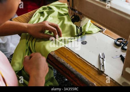 Close up of asian seamstress hands guiding green fabric through a sewing machine. Essential tools like scissors and a seam ripper are nearby, highligh Stock Photo