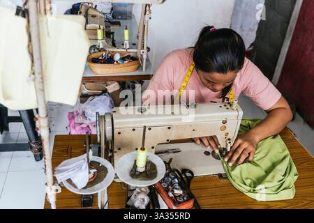 A asian seamstress focuses intently on sewing fabric using a vintage machine. The workshop is filled with tools, threads, and measuring tape, highligh Stock Photo