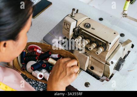 A asian seamstress selects colorful threads from a basket beside an industrial sewing machine, showcasing the art of tailoring and garment creation in Stock Photo