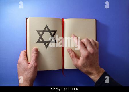 Concept of a religious Jew reading from an open book with a religious symbol, holding the book with one hand and pointing with the other on a blue bac Stock Photo