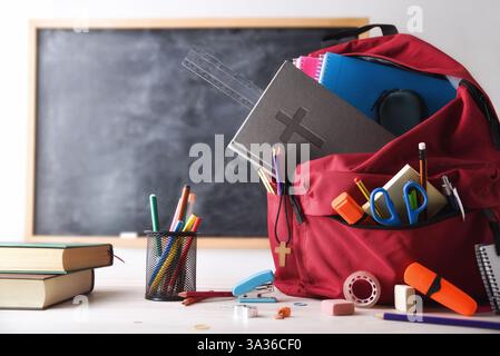 School education in a religious school with a red backpack full of tools and school supplies and a Bible on a desk with books and supplies in a classr Stock Photo
