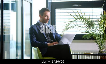 Business professional working on laptop in modern office space with sunlight streaming through large windows. Stock Photo