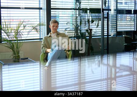Young professional dressed casually, working on laptop in modern office space with sunlight streaming through blinds. Stock Photo