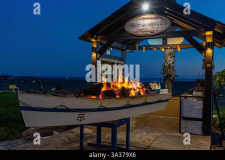 A nautical-themed barbeque featuring a boat filled with burning coals, ready for grilling, in an outdoor setting at dusk. Stock Photo