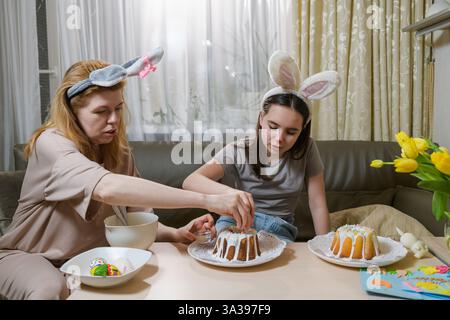 A mother and daughter wearing bunny ears are decorating Easter cakes with white icing and colorful sprinkles. They sit at a table with painted eggs, f Stock Photo