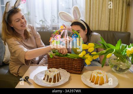 A mother and daughter wearing bunny ears are joyfully decorating an Easter basket with yellow tulips, handmade cards, and festive ornaments. Two glaze Stock Photo