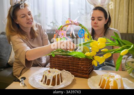 A mother and daughter wearing bunny ears are joyfully decorating an Easter basket with yellow tulips, handmade cards, and festive ornaments. Two glaze Stock Photo