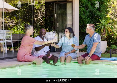 Senior Friends enjoying drinks by poolside, laughing and relaxing in sunny backyard Stock Photo