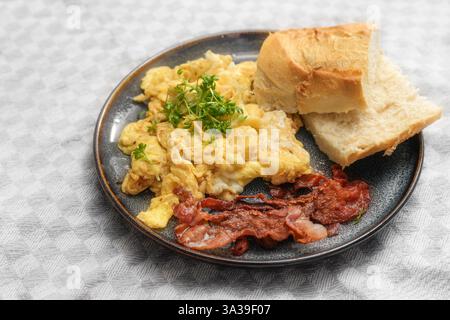 Scrambled eggs with crispy bacon, bread and cress garnish on a plate and a light gray tablecloth, hearty breakfast or quick dinner, selected focus, na Stock Photo