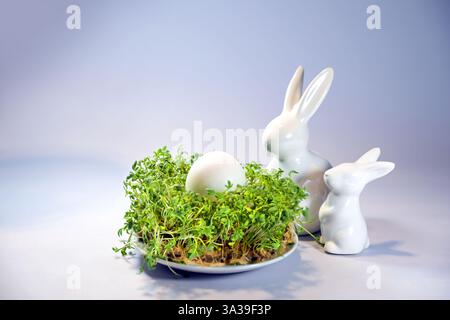Two Easter bunnies from white ceramic looking to an egg placed in a bed of cress on a plate, light purple blue background, copy space, selected focus, Stock Photo