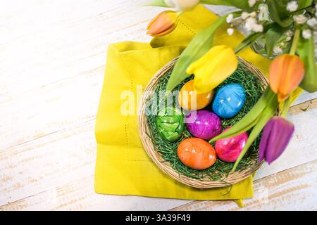 Colorful painted Easter eggs in a basket and a spring bouquet with tulips on a yellow napkin and a white wooden background, top view from above, copy Stock Photo