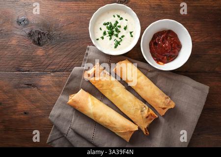 Crispy baked filo pastry rolls or spring rolls with two dips in white bowls on a gray napkin and a dark rustic wooden table, homemade finger food, top Stock Photo