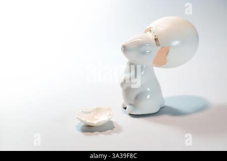 White ceramic Easter bunny wears an empty eggshell like a hat on his head, funny seasonal greeting card, light background, copy space, selected focus, Stock Photo