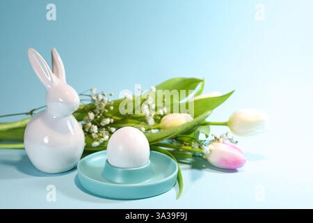 White Easter bunny from ceramic, an egg in a turquoise cup and a spring flower bouquet with rose tulips against a turquoise background, copy space, se Stock Photo