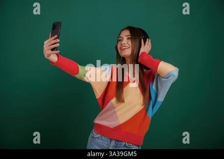 Making selfie. Smiling, in headphones. Young woman is standing against green background in the studio. Stock Photo