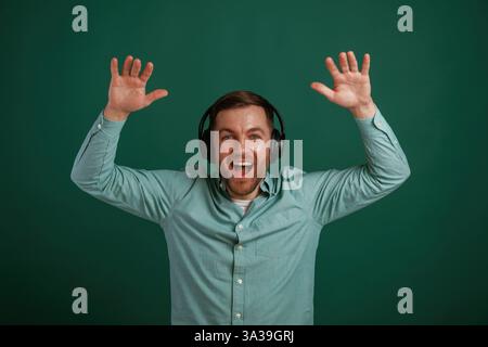 With hands up, in headphones. Man is standing against background in the studio. Stock Photo