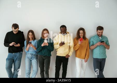 Busy by using smartphones. Group of young people are standing against white background. Stock Photo