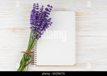 Small bouquet of fresh blooming lavender lies on blank spiral paper notebook, notepad on white wooden background. Mockup, template with copy space, to Stock Photo
