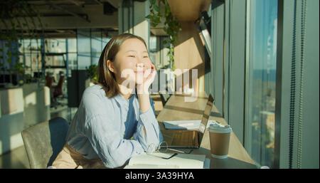 A young woman in a bright modern office, enjoying a calm moment at her desk with a laptop, notebook, and coffee, surrounded by greenery and natural Stock Photo